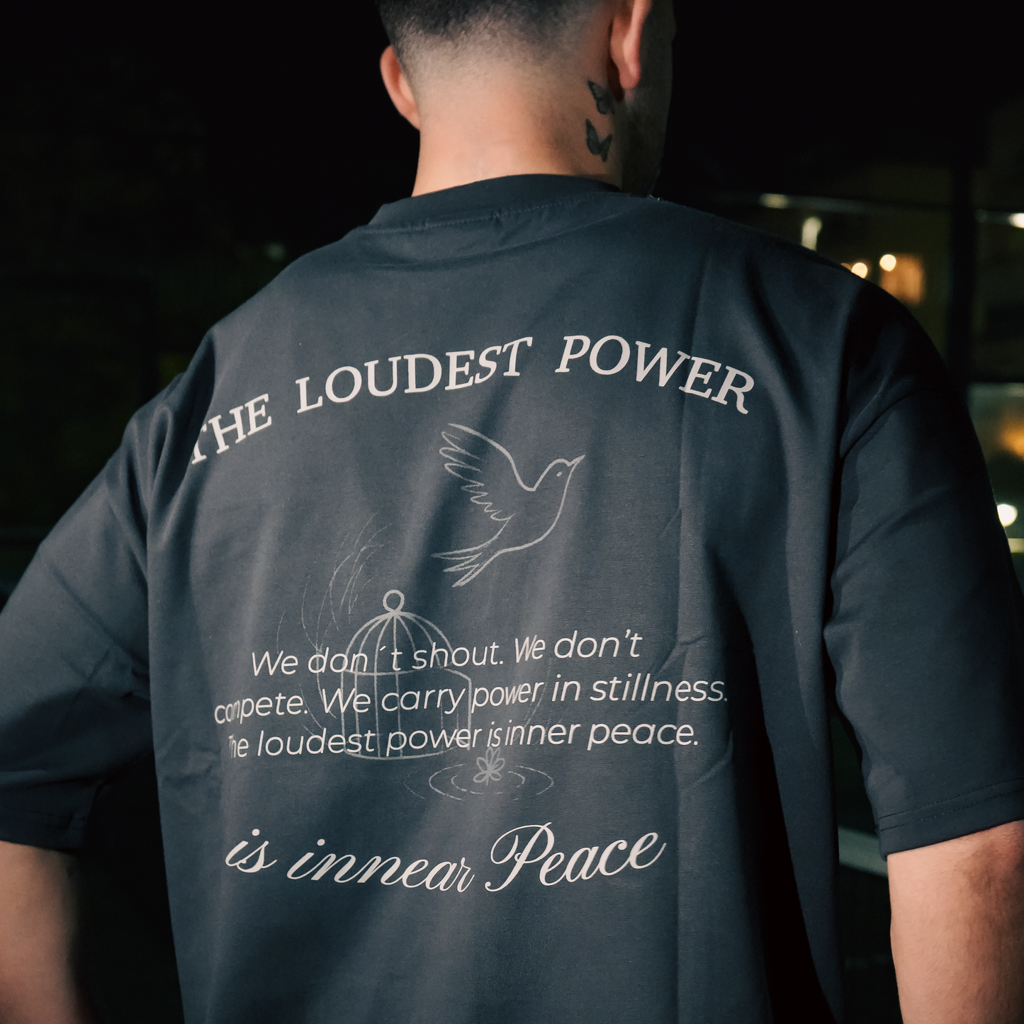 Person wearing a black t-shirt with text and a dove graphic on the back, standing against a dark background.